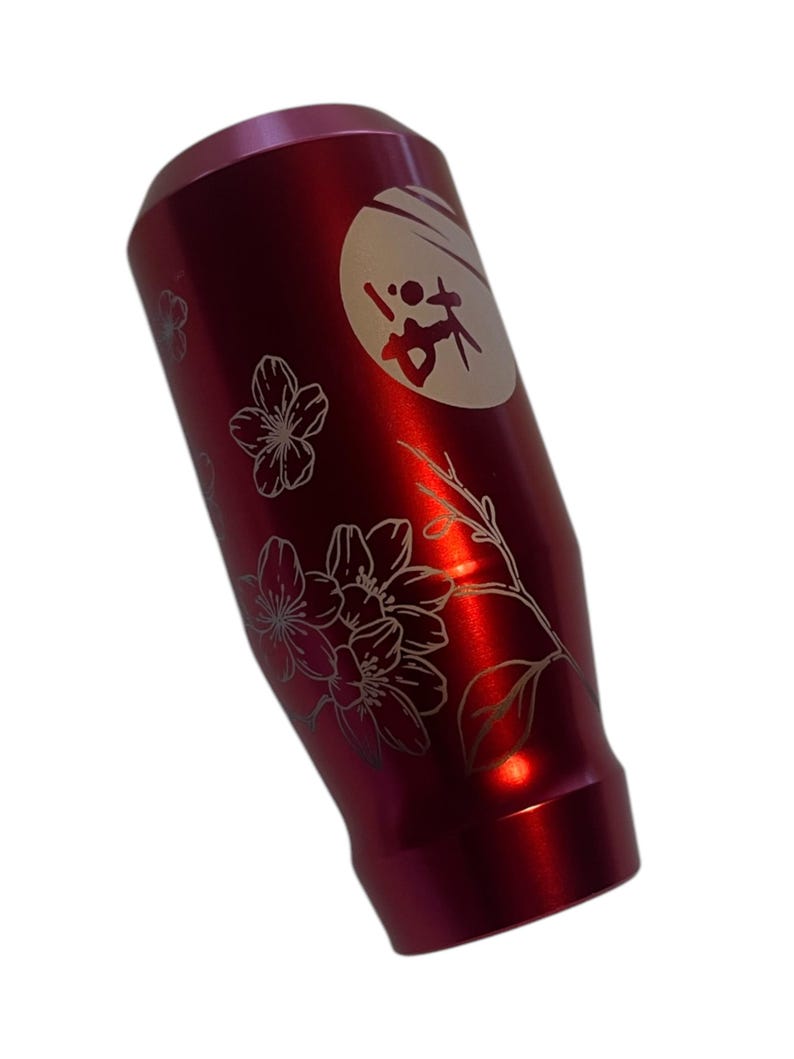 Pu&ograve; includere: Pomello del cambio in alluminio rosso con un design inciso bianco di fiori di ciliegio e un cerchio con caratteri giapponesi.