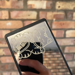 Puede incluir: Un espejo compacto rectangular con borde negro. El espejo presenta un diseño grabado de un gato de dibujos animados con los caracteres chinos de "riqueza" o "fortuna". El fondo es una pared de ladrillos.