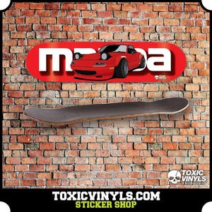 Op de afbeelding: Een rode Mazda Miata auto sticker op een zwart skateboard deck. De sticker is op een rode achtergrond met witte tekst die "Mazda" zegt. Het skateboard hangt aan een bakstenen muur. De tekst "TOXIC VINYLS.COM STICKER SHOP" staat onderaan de afbeelding.