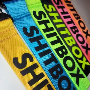 Pode incluir: Um conjunto de seis cordões coloridos com a palavra "SHITBOX" impressa em preto em cada um. Os cordões são amarelos, azuis, verdes, pretos, rosas e laranjas.