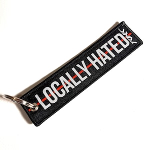 Pode incluir: Chaveiro preto com uma faixa vermelha e texto branco que diz "LOCALLY-HATED".