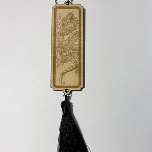 May include: A wooden bookmark with a laser-engraved image of a dragon. The bookmark has a black tassel attached to it.