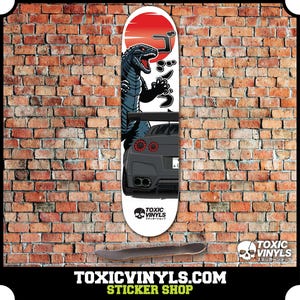 May include: A white skateboard deck with a black and gray illustration of a Godzilla-like monster and a car. The monster is standing on its hind legs with its mouth open and its arms raised. The car is a black sports car with red taillights. The background is a red sun with a white circle around it. The text "TOXIC VINYLS" is printed on the bottom of the deck.