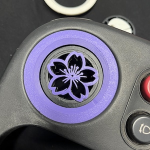 May include: A black video game controller featuring a custom purple analogue stick cap shaped like a cherry blossom flower. The controller includes a red button and a black button, with a white circular ring component visible in the background.