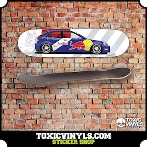 Könnte beinhalten: Ein Skateboard-Deck mit einem Grafikdruck eines blauen und roten Rennwagens mit dem Text "NO-GOOD.com" und "Red Bull" auf weißem Hintergrund. Das Skateboard hängt an einer Backsteinwand.
