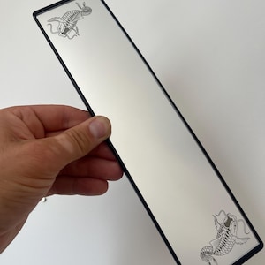 May include: A long, rectangular mirror with a thin black frame. The mirror surface features two black line-art illustrations of koi fish, one positioned in the top left corner and the other in the bottom right corner of the reflective glass.