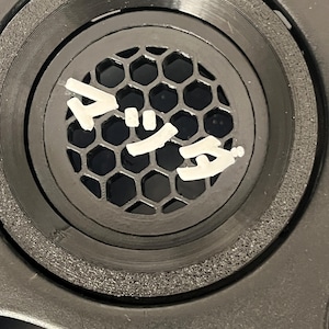 May include: A close-up shot of a black circular component featuring a honeycomb mesh. The component has concentric rings and a textured outer edge. White Japanese characters are visible on the surface.