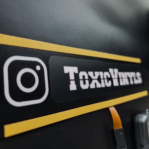 Pode incluir: Um adesivo preto e amarelo com um logotipo do Instagram branco e o texto "ToxicVinyls" em letras brancas.