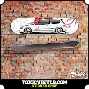 Könnte beinhalten: Ein weißer Honda Civic mit einem roten und weißen Kirschblüten-Design auf einem Skateboard-Deck. Das Skateboard-Deck hängt an einer Backsteinwand. Der Text "TOXIC VINYLS" befindet sich auf dem Skateboard-Deck.