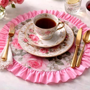 Può includere: Un servizio da tè floreale con un sottobicchiere rosa a balze. Il set include una tazza, un piattino e un piatto, tutti decorati con rose rosa e bordi dorati. Posate dorate e un tovagliolo di pizzo completano l'elegante mise en place.