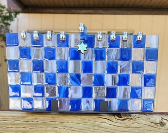 May include: A rectangular glass menorah featuring a blue and white checkered pattern. The piece has nine silver-toned candle holders along the top edge and a central hanging Star of David charm. The structure is supported by two black metal brackets.