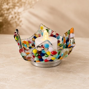 May include: A glass candle holder with a wavy, flared rim, constructed from numerous small, multicolored glass beads fused together. A white, battery-operated tea light candle with a warm, glowing flame sits in the center of the bowl.