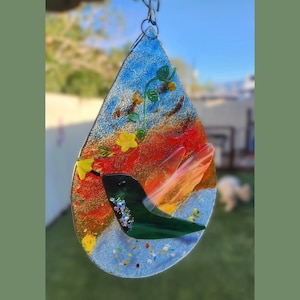 May include: A teardrop-shaped fused glass suncatcher featuring a dark green hummingbird in flight. The background displays a gradient of blue, orange, and red glass textures. A green vine with small yellow flowers curves across the upper section.