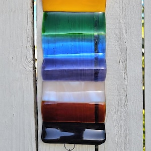 May include: A rectangular fused glass wall hanging featuring horizontal bands of deep red, bright yellow, green, blue, purple, white, brown, and black. A small metal chain hangs from the bottom, attached to a clear glass triangle with a light blue heart.