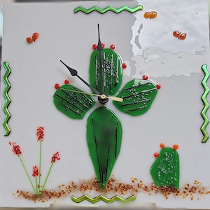 May include: A white square clock with a green glass cactus design. The clock has black hands and a gold second hand. The cactus is surrounded by small red and brown glass pieces. The clock has a green and gold zig-zag border.