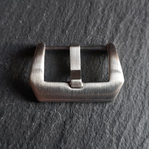 Puede incluir: Una hebilla de correa de reloj plateada con acabado cepillado.