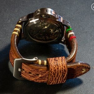 Puede incluir: Una correa de reloj de cuero marr&oacute;n con costuras blancas y una hebilla de metal. La correa tiene una franja verde, blanca y roja en la parte superior.