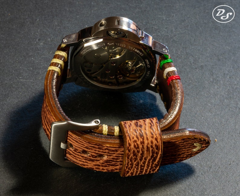 Puede incluir: Una correa de reloj de cuero marr&oacute;n con costuras blancas y una hebilla de metal. La correa tiene una franja verde, blanca y roja en la parte superior.