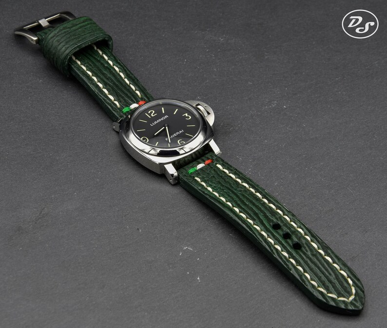 May include: A green leather watch strap with white stitching and a green, white, and red accent. The strap is attached to a silver watch with a black face and white markings.
