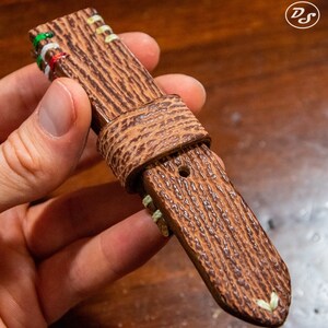 Puede incluir: Una correa de reloj de cuero marr&oacute;n con costuras blancas y una superficie texturizada. La correa tiene un agujero para una hebilla y est&aacute; doblada en la parte superior.