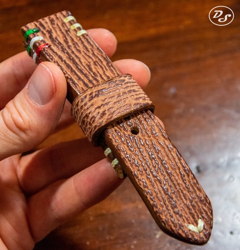 Puede incluir: Una correa de reloj de cuero marr&oacute;n con costuras blancas y una superficie texturizada. La correa tiene un agujero para una hebilla y est&aacute; doblada en la parte superior.