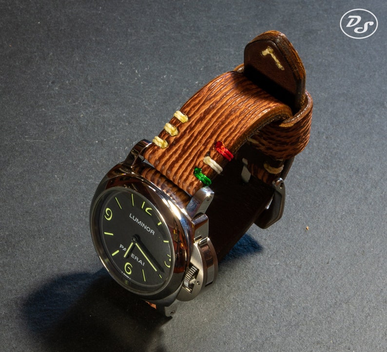 Puede incluir: Una correa de reloj de cuero marr&oacute;n con costuras blancas y detalles coloridos. La correa est&aacute; unida a un reloj plateado con una esfera negra y la palabra "Luminor" impresa en ella.