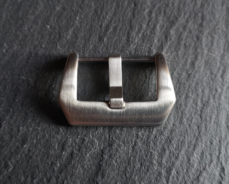 May include: A silver watch band buckle with a brushed finish.