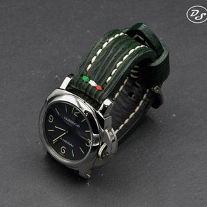 May include: A black dial watch with white markings and the words "Luminor" and "Panerai" on the face. The watch has a green leather strap with white stitching and a small Italian flag detail.