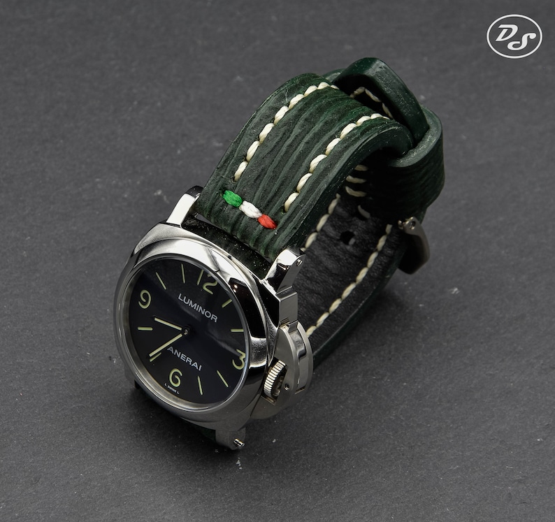 May include: A black dial watch with white markings and the words "Luminor" and "Panerai" on the face. The watch has a green leather strap with white stitching and a small Italian flag detail.
