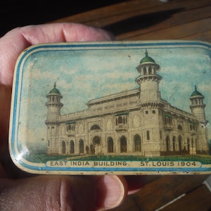Könnte beinhalten: Eine Vintage-Blechdose mit blau-weißem Rand. Die Dose zeigt eine farbenfrohe Illustration des East India Building in St. Louis, Missouri, erbaut im Jahr 1904. Der Text "East India Building, St. Louis 1904" ist auf der Unterseite der Dose aufgedruckt.