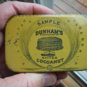 May include: A yellow metal tin with black lettering that reads "Sample Dunham's Concentrated Shred Cocoanut" with a black and white illustration of a cake on top.