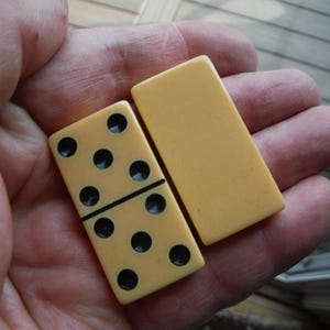 May include: Two ivory-colored dominoes. One domino shows seven black dots, and the other is blank. The dominoes are rectangular and have rounded corners. The dominoes are held in a hand.