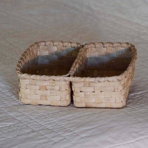 May include: A woven basket with two compartments. The basket is made of light brown, natural-colored reeds. The rectangular basket has a woven design and is suitable for storage or display.