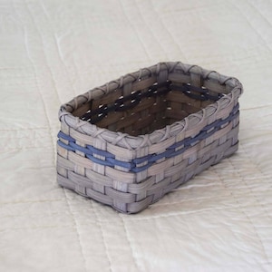 May include: A woven basket with a rectangular shape and a light gray color. The basket has a blue trim around the top and bottom.