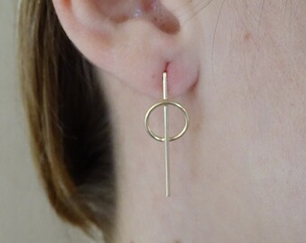 May include: A pair of gold-colored earrings. Each earring features a small, delicate circle suspended from a vertical bar. The earrings are simple and modern in design, with a minimalist aesthetic. The earrings are shown on a person's ear.