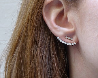 May include: A close-up of an ear adorned with two pieces of jewelry. One is a small, delicate stud with three square-shaped stones. The other is a longer, ear-climbing earring with a series of square-shaped stones, creating a sparkling, geometric design.