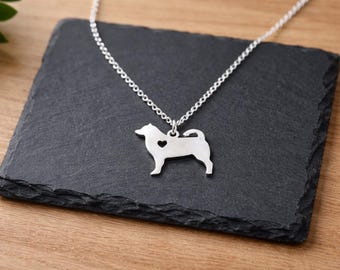 May include: A silver-colored dog pendant necklace with a heart-shaped cutout on a chain. The dog pendant is displayed on a dark gray, textured surface. The background is a wooden surface.