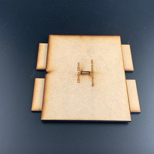 以下が含まれることがあります： ミニチュアウォーゲームの地形用パーツの茶色の木製ベース。ベースは125mm&times;100mmで、中央に小さなスロットがあります。ベースには「WarchestCreations.com」の文字が印刷されています。
