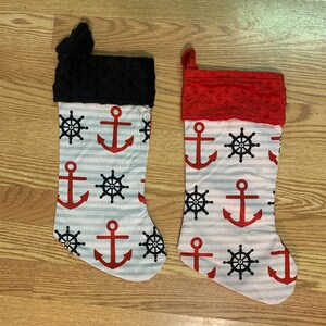 May include: Two nautical-themed Christmas stockings. One has a black cuff, the other a red cuff. Both have a white and gray striped pattern with red anchors and black ship wheels. The stockings are made of fabric.