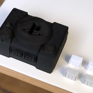 May include: A black 3D printed PlayStation console with a PlayStation logo on the top. The console has multiple ports on the front. There are four white 3D printed controllers on a white surface.