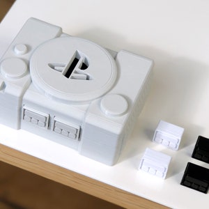 May include: A white 3D printed model of a PlayStation console with a PlayStation logo on the top. There are four controller buttons, two white and two black, next to the console.