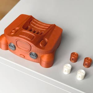 May include: A 3D printed orange Nintendo 64 controller with four grey buttons and a black cartridge slot. The controller is sitting on a white surface with four small orange and white buttons.