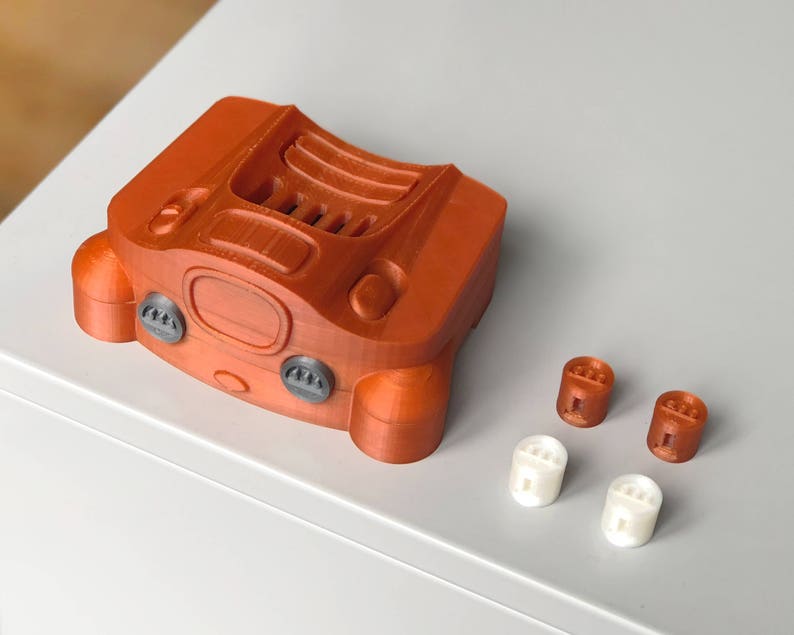 May include: A 3D printed orange Nintendo 64 controller with four grey buttons and a black cartridge slot. The controller is sitting on a white surface with four small orange and white buttons.
