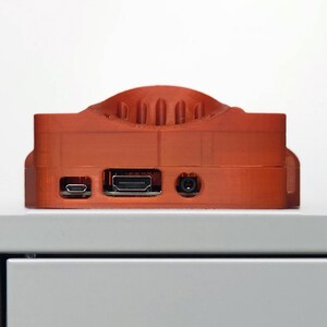 May include: An orange 3D printed case for a Raspberry Pi computer. The case has a raised section on the top and includes ports for HDMI, USB, and power.