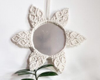 May include: A white macrame mirror with a round mirror in the center, hanging on a white wall with a green plant in the foreground.