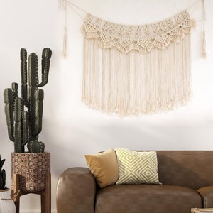 May include: A brown leather sofa with two pillows, one yellow and one with a white and yellow geometric pattern. A large cactus plant in a woven basket sits on a wooden stand in the corner of the room. A macrame wall hanging with a fringe design hangs above the sofa.