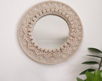 May include: A round mirror with a macrame frame made of natural beige cotton cord. The mirror is hanging on a white wall with a green plant in the foreground.