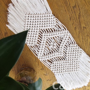 May include: A white macrame table runner with a geometric pattern and fringe edges. The runner is on a wooden surface.