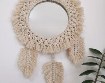 May include: A round mirror with a macrame frame and fringe, decorated with wooden beads. Three macrame feathers hang from the bottom of the mirror.