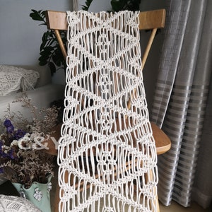 May include: A white macrame table runner with a geometric pattern and fringe. The runner is draped over a wooden chair.